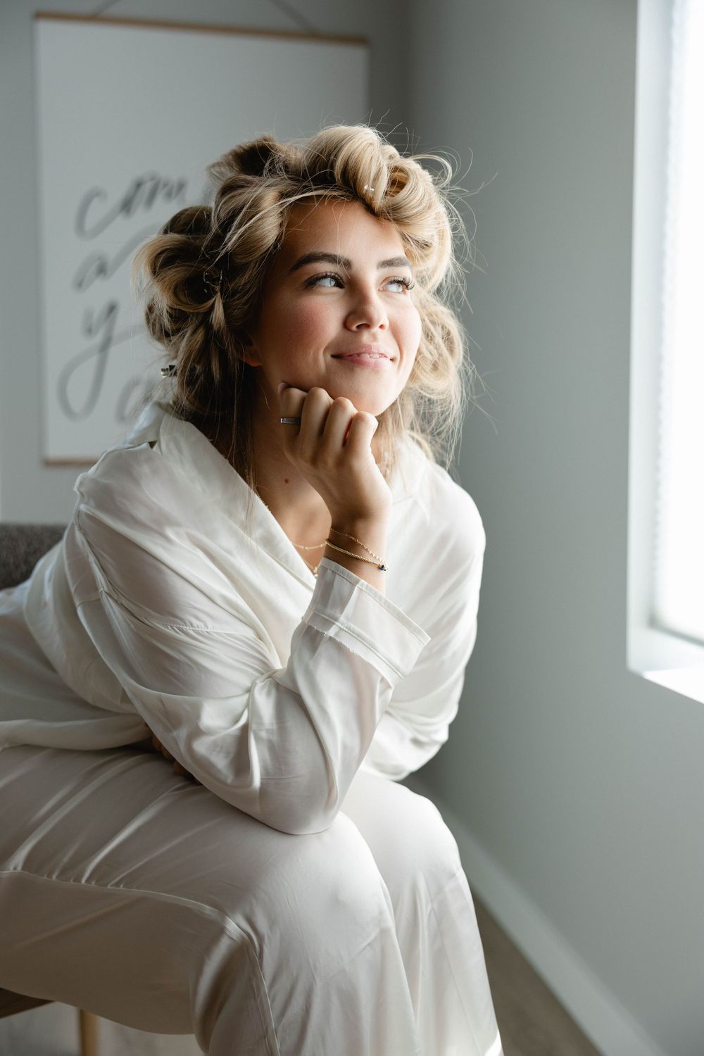 Person with blonde curled hair sits by window in white robe, resting chin on hand in contemplative pose.