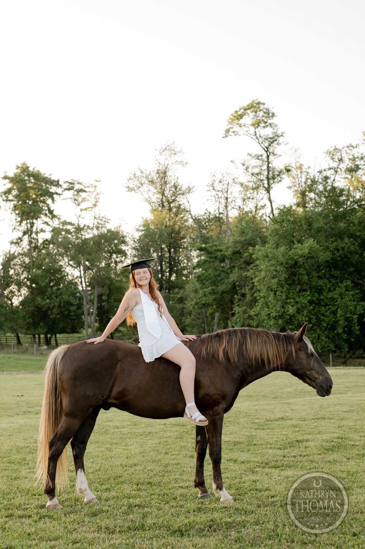 A graduate in white dress sits bareback on a dark horse in a pastoral setting with trees.