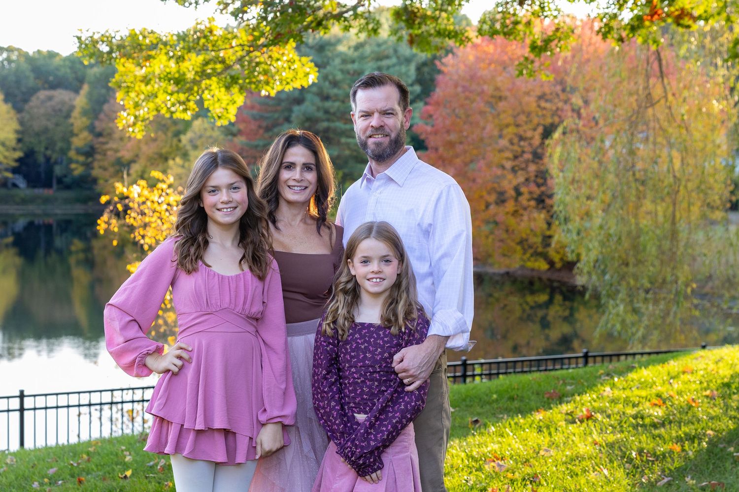 A family poses together by a serene pond with colorful fall foliage in the background during a family photo session in Massachusetts.