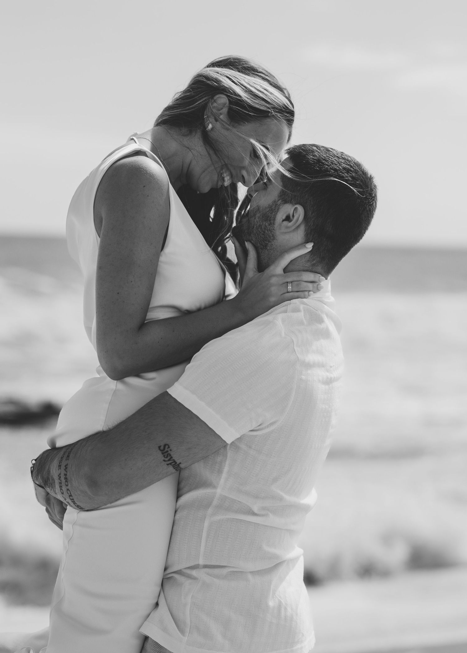 A romantic black and white beach photo of a couple embracing, with the ocean visible in the background.