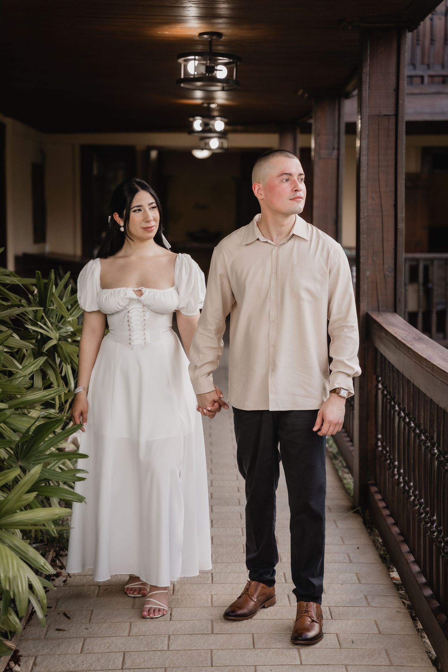 Couple walking hand in hand along a covered walkway, with woman in white dress and man in beige shirt.