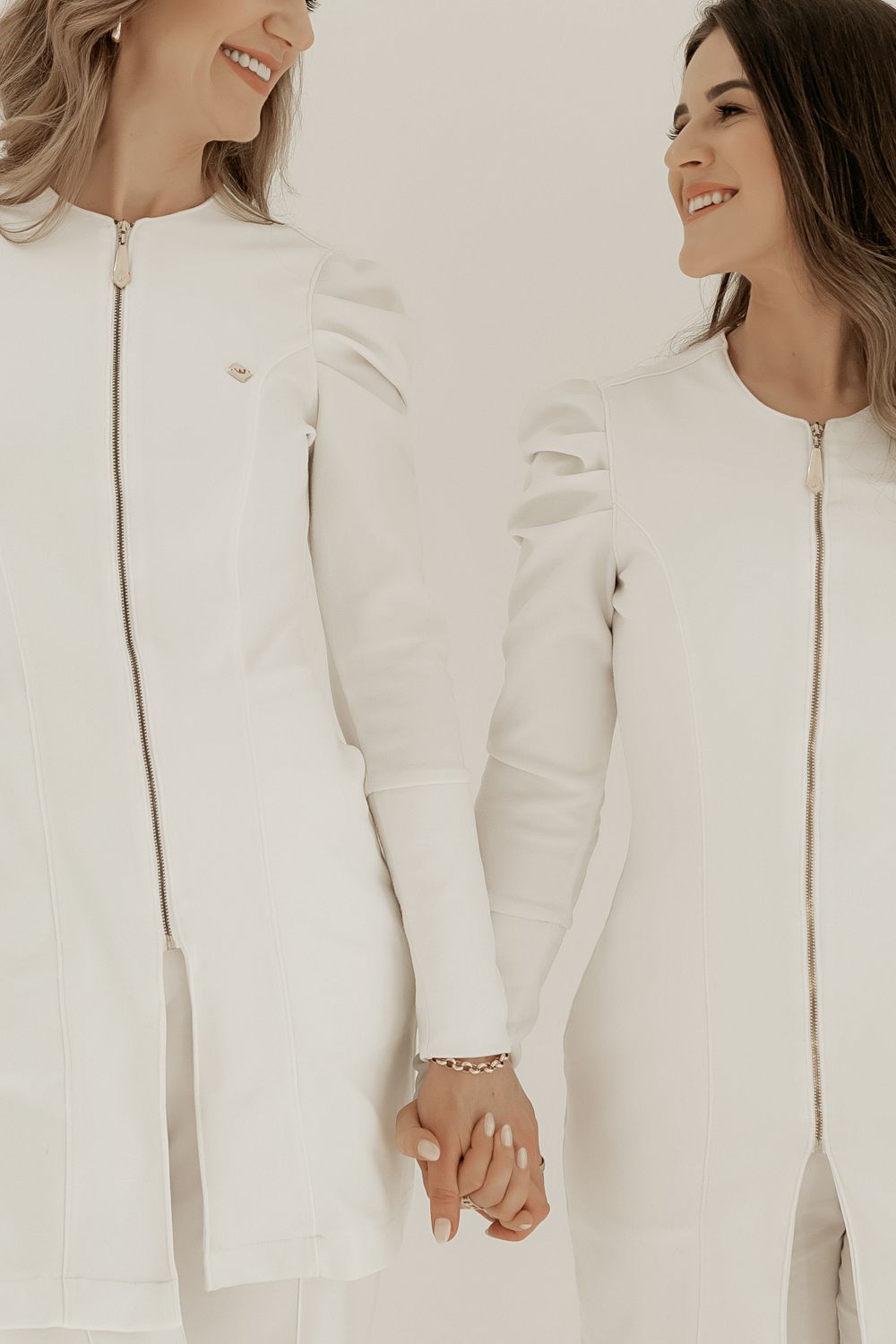 Two people wearing matching white zip-up outfits hold hands while smiling in a minimalist portrait setting.