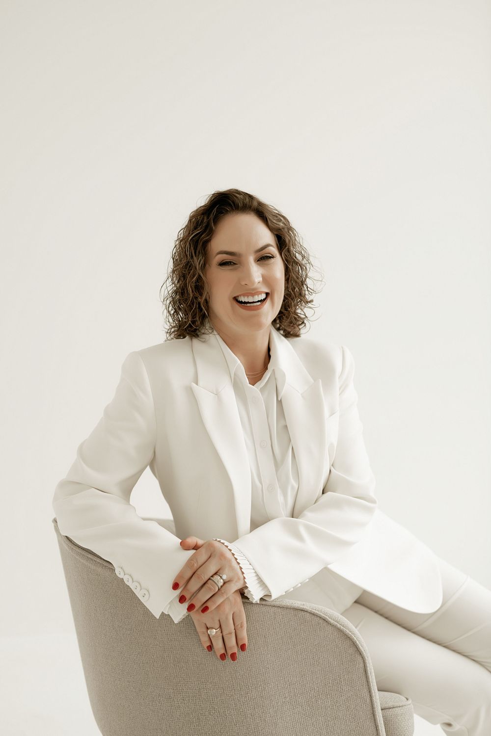 Professional wearing an elegant white suit strikes a relaxed pose while seated in a light gray chair.
