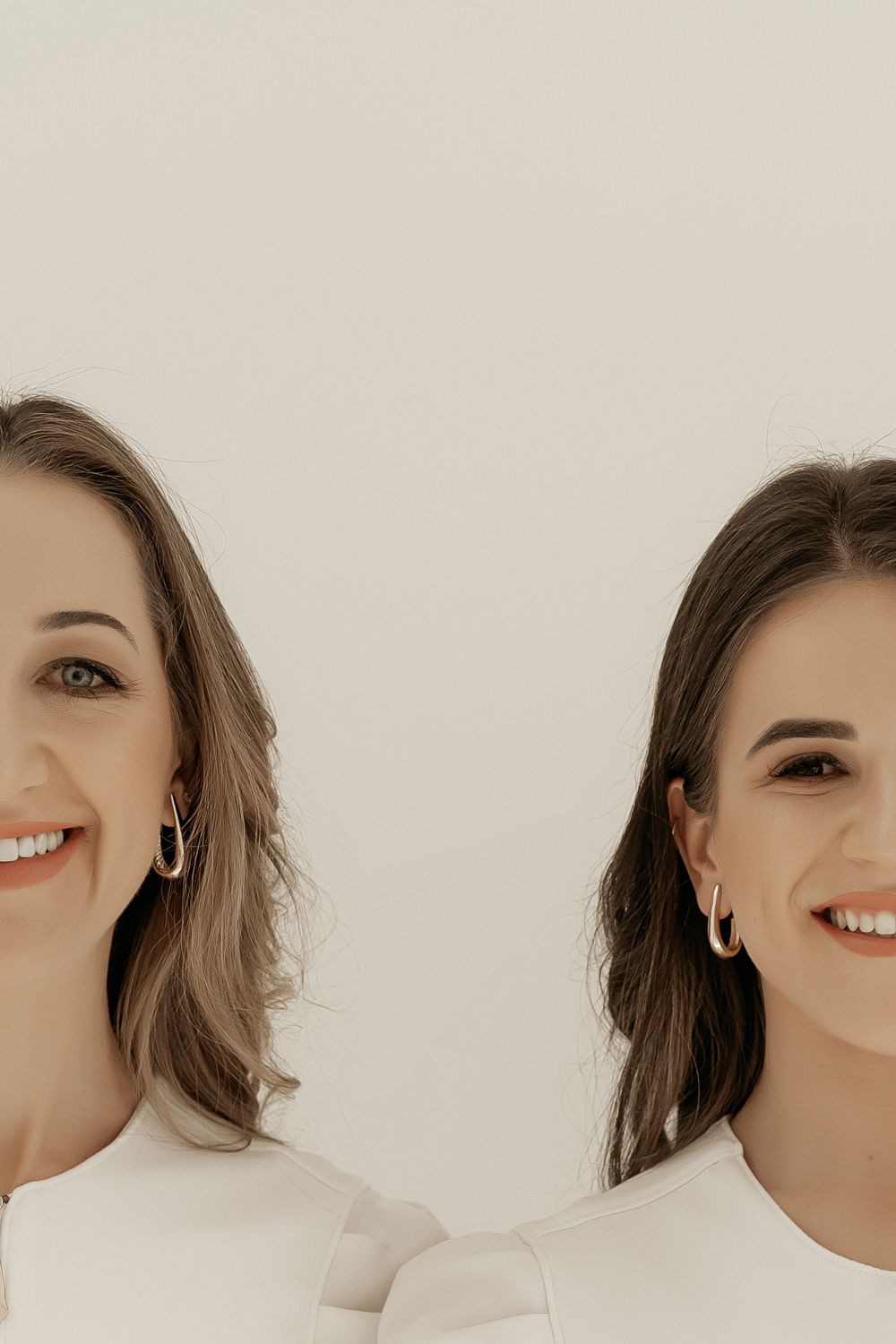 Two women with bright smiles and brown hair wearing white outfits against a neutral background.