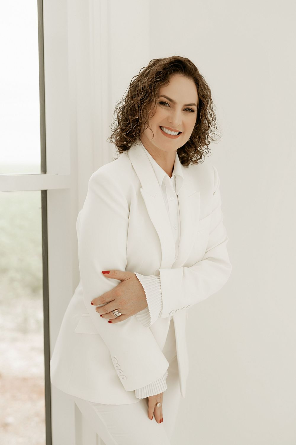Professional portrait of someone wearing an elegant all-white suit with natural curly hair against a light background.