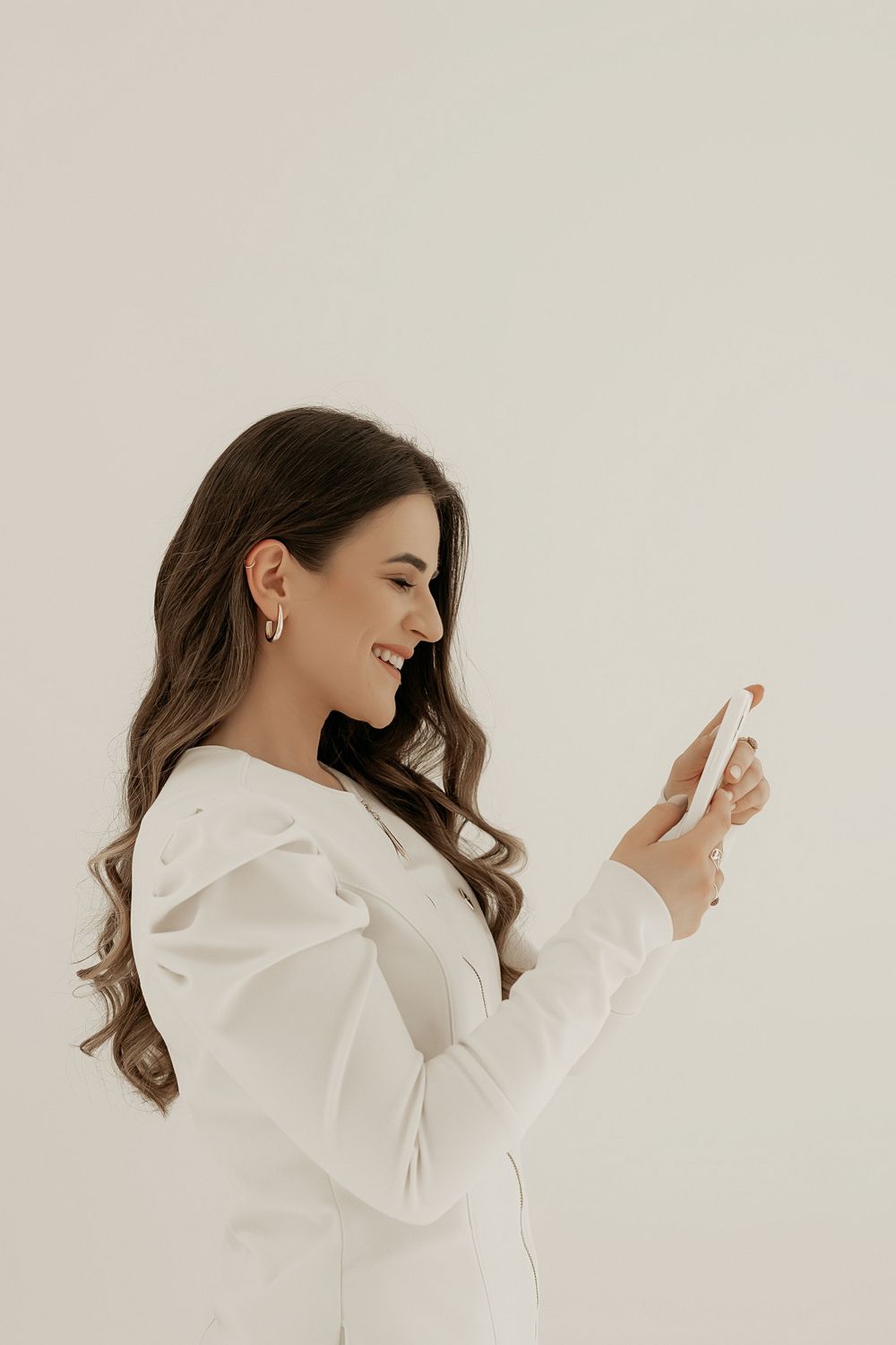 Side profile of person with long wavy brown hair wearing white business attire using a smartphone against light background.