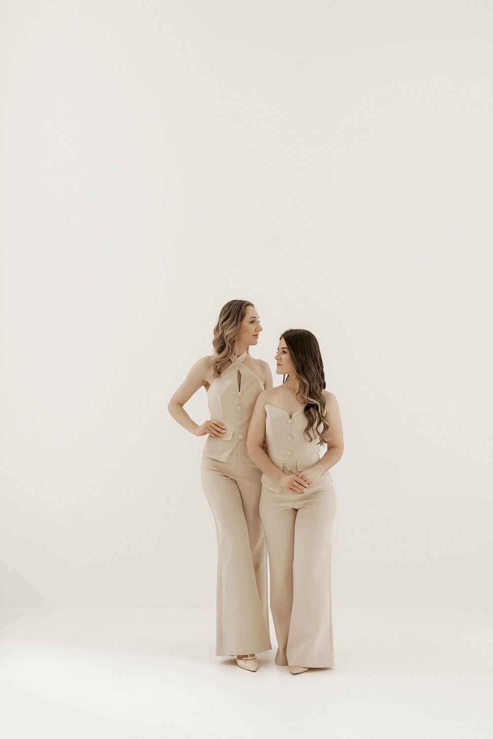 Two people wearing matching beige outfits stand together against a white backdrop in a minimalist studio setting.
