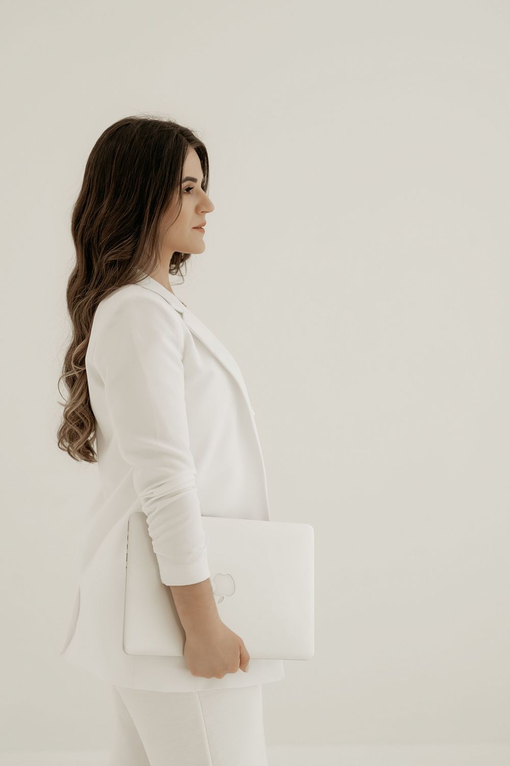 Person wearing an all-white business outfit holding a laptop while standing against a light background in a minimalist setting.