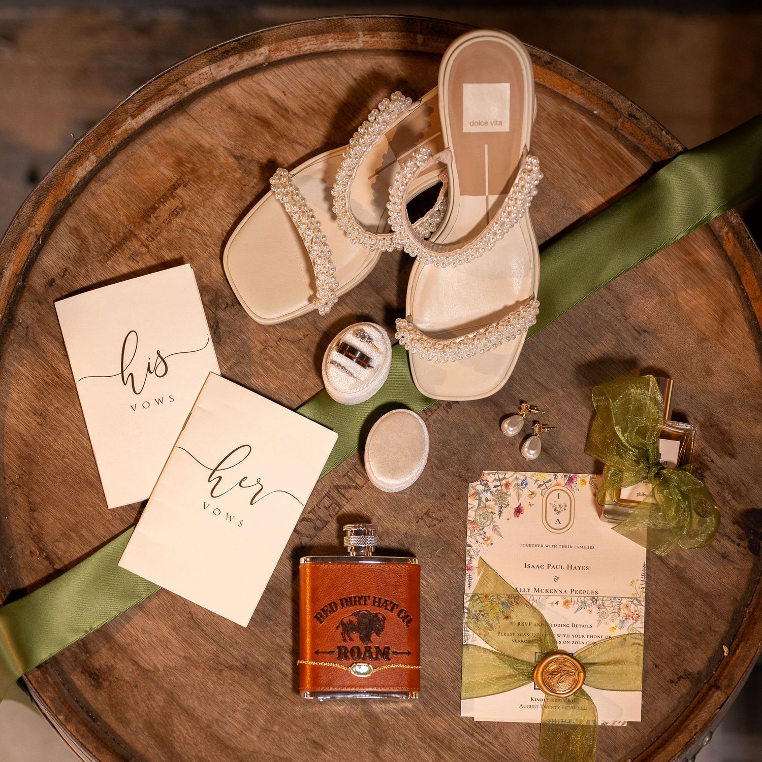 Wedding accessories including beige sandals, stationery, green ribbon, and a bottle of bourbon arranged on a wooden surface.