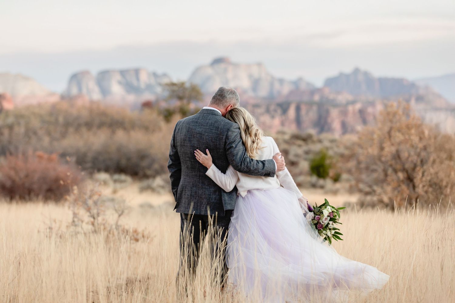 A couple in wedding attire embraces while looking at scenic mountain views across desert terrain and tall grass.