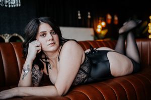 Person in black lingerie poses on brown leather couch in dimly lit setting.