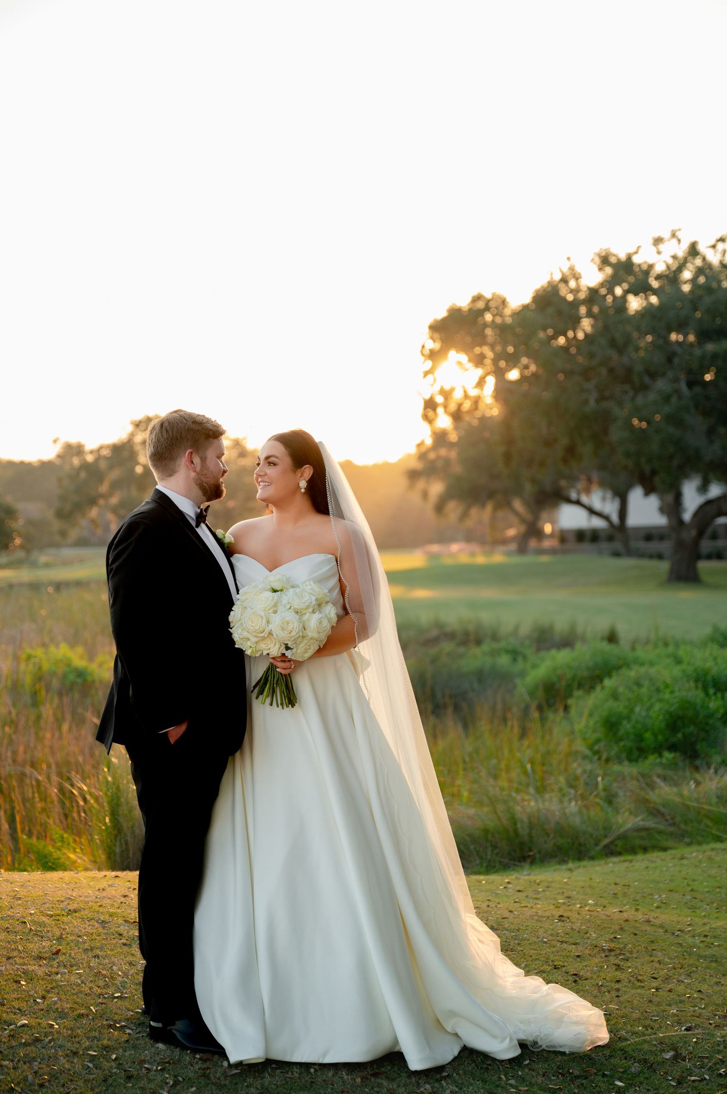 Wedding couple poses in elegant attire against sunset landscape with flowing veil.