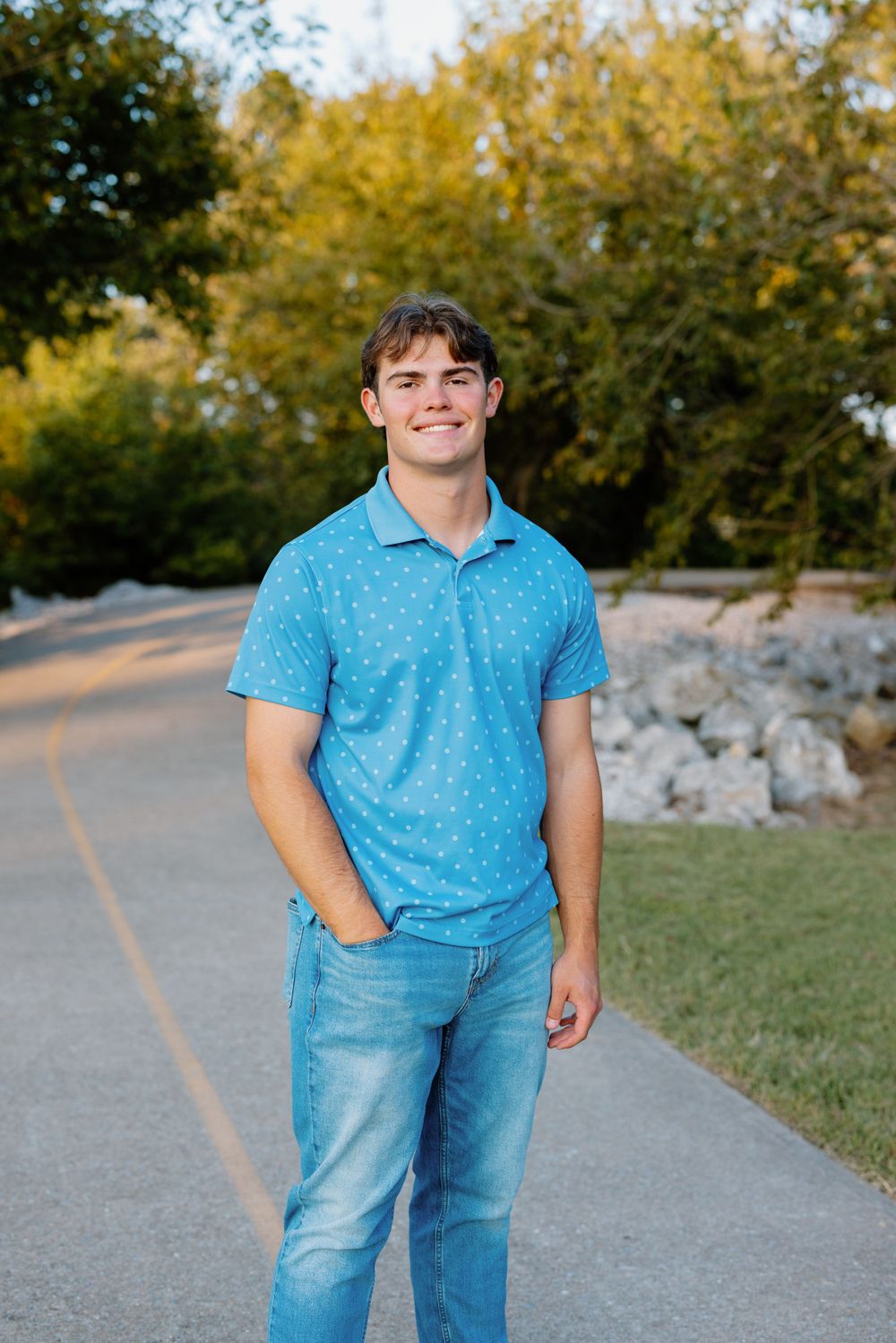 Person in blue polo shirt and jeans stands on outdoor path with autumn trees in background.