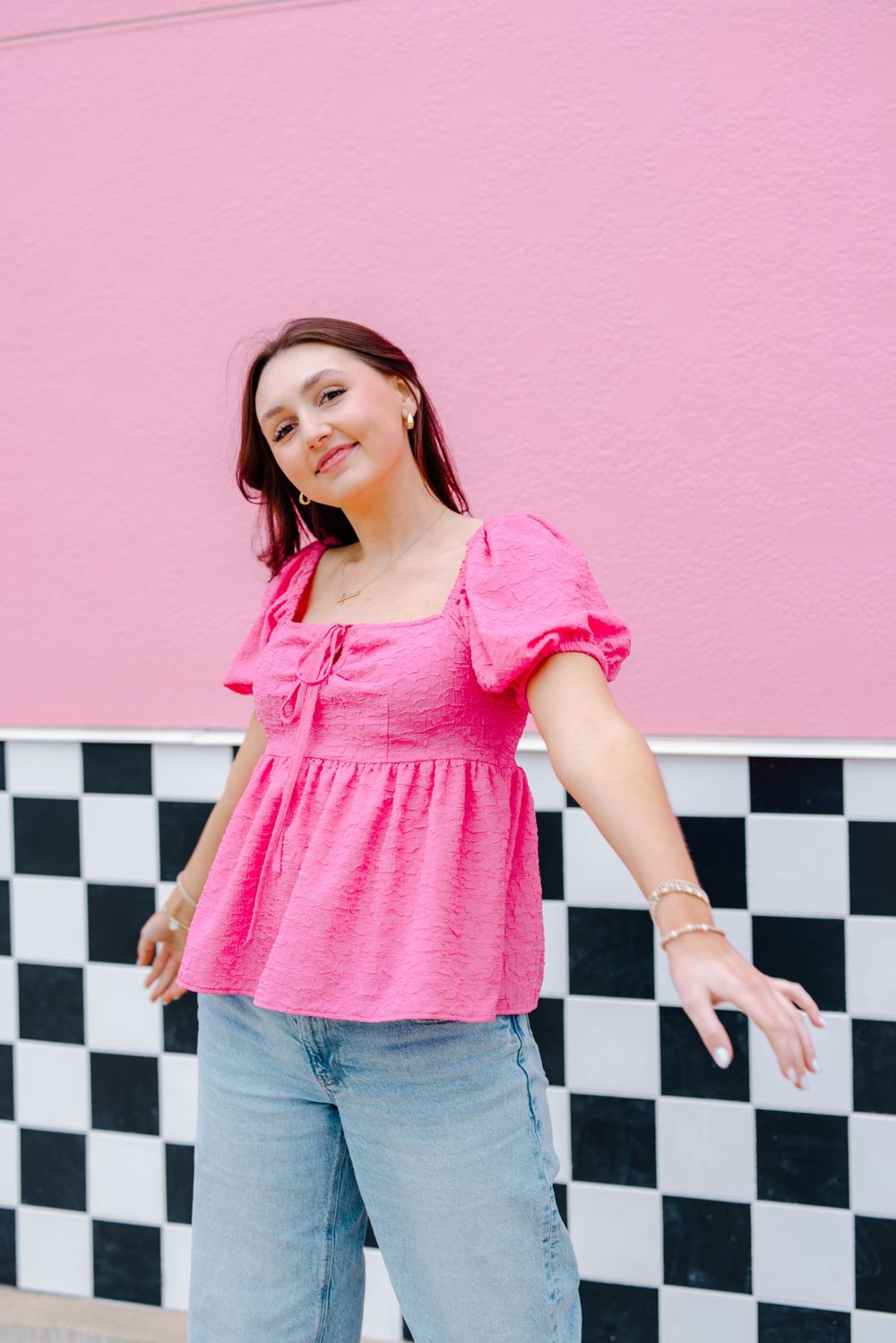 A bright pink puff sleeve blouse is styled with blue jeans against a pink wall with checkerboard trim.