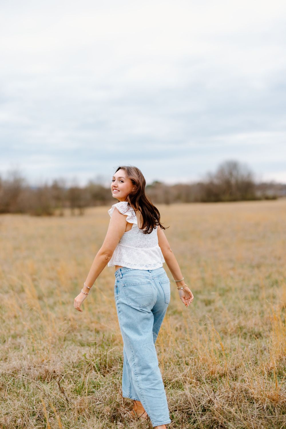 A person in light denim jeans and white top twirls joyfully in a golden autumn field under cloudy skies.