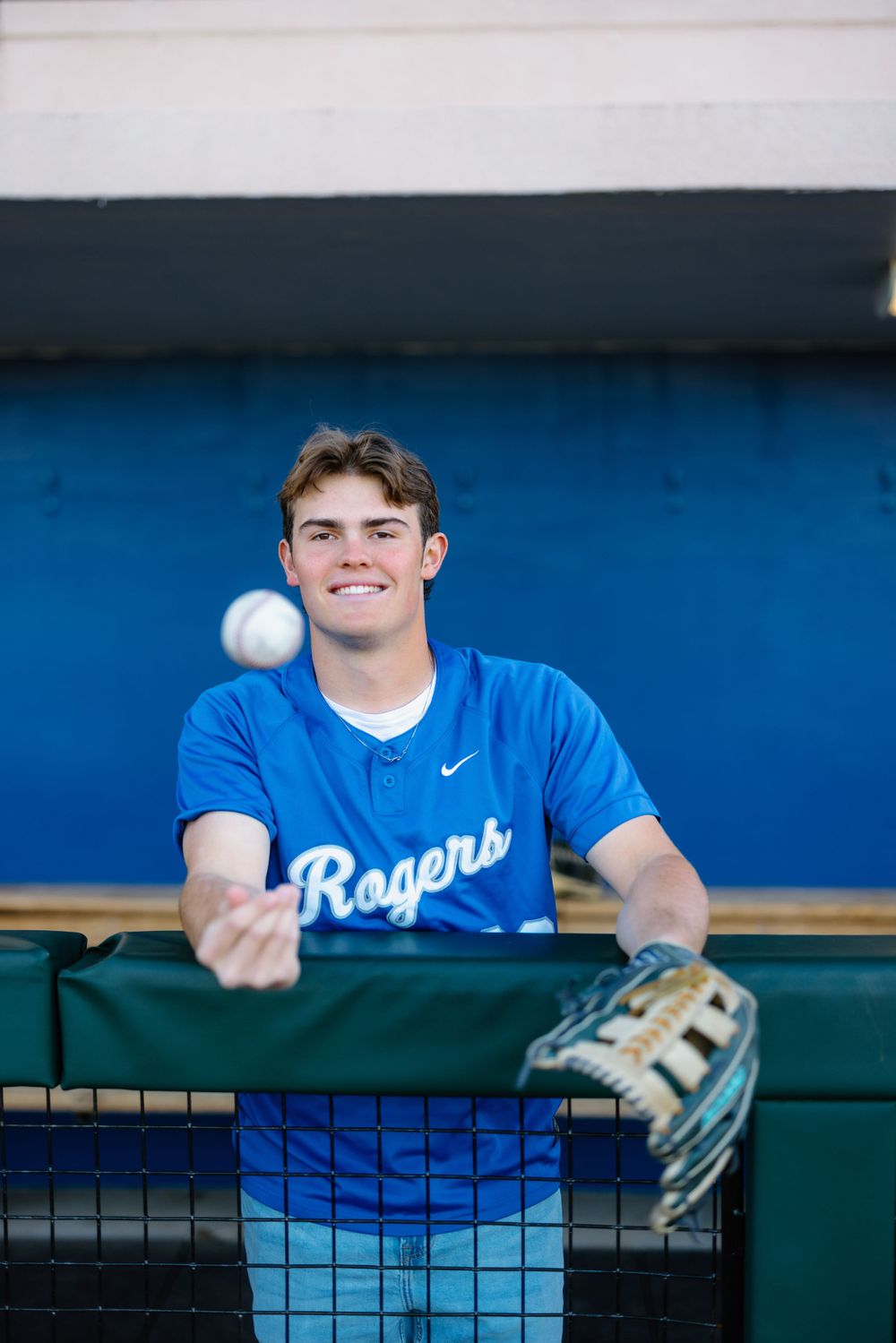 Baseball player in blue Rogers jersey smiles while leaning on dugout fence with baseball and glove.