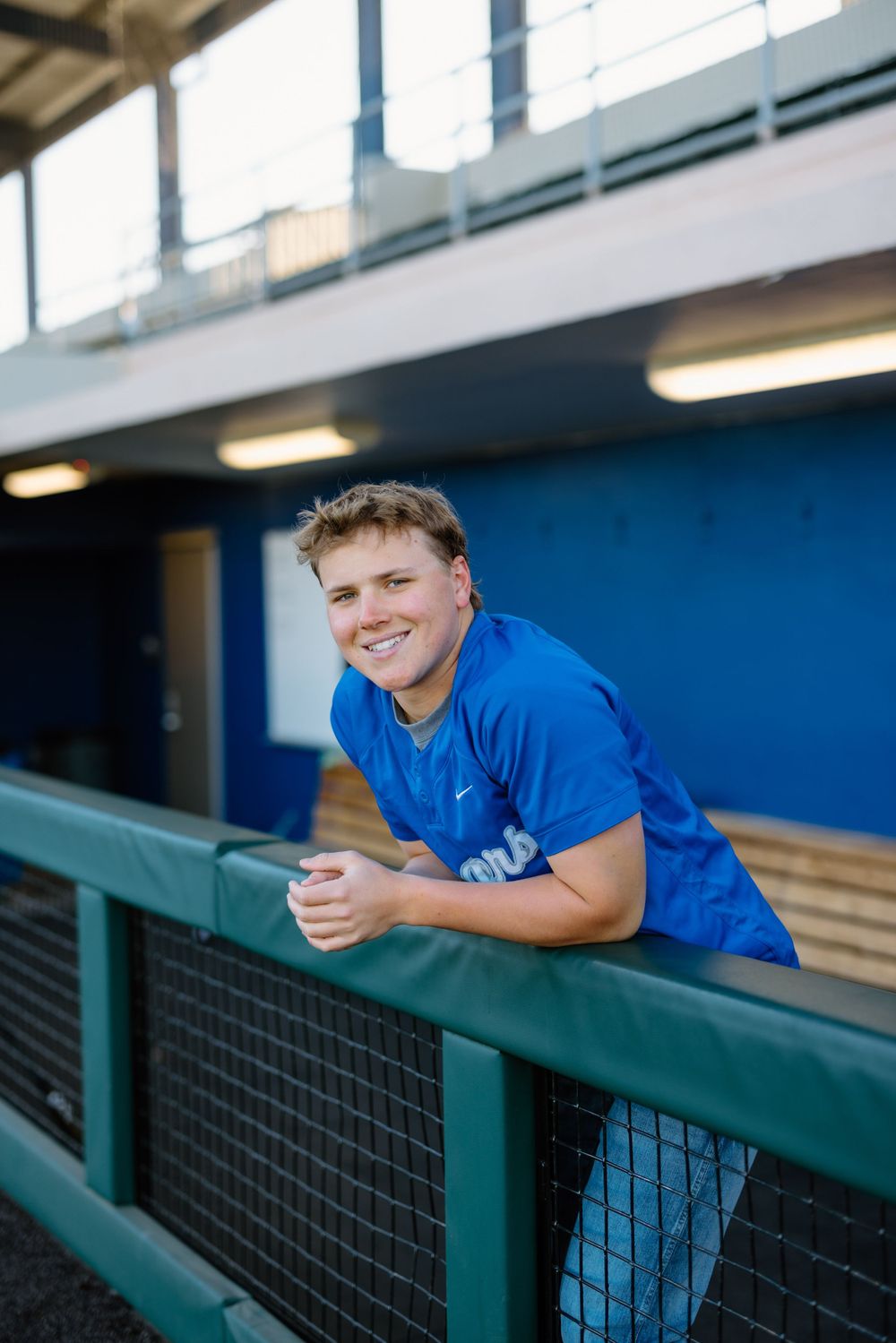 Baseball player in blue uniform leans against dugout railing with a friendly expression.
