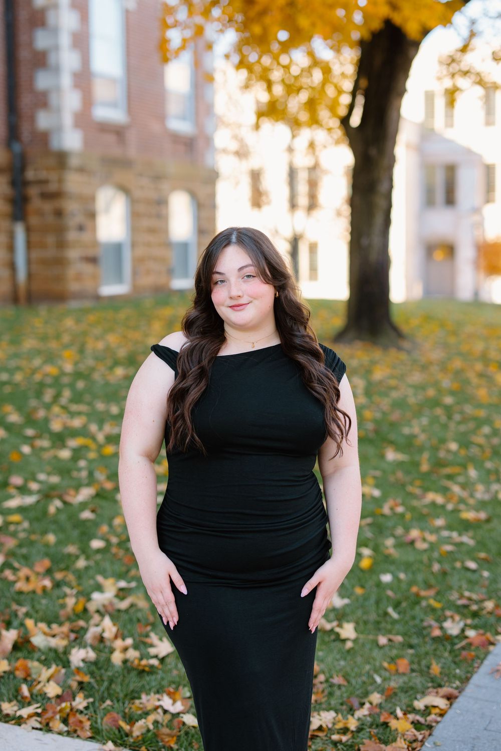 Young woman in black dress poses outdoors among autumn leaves with golden foliage and brick building backdrop.