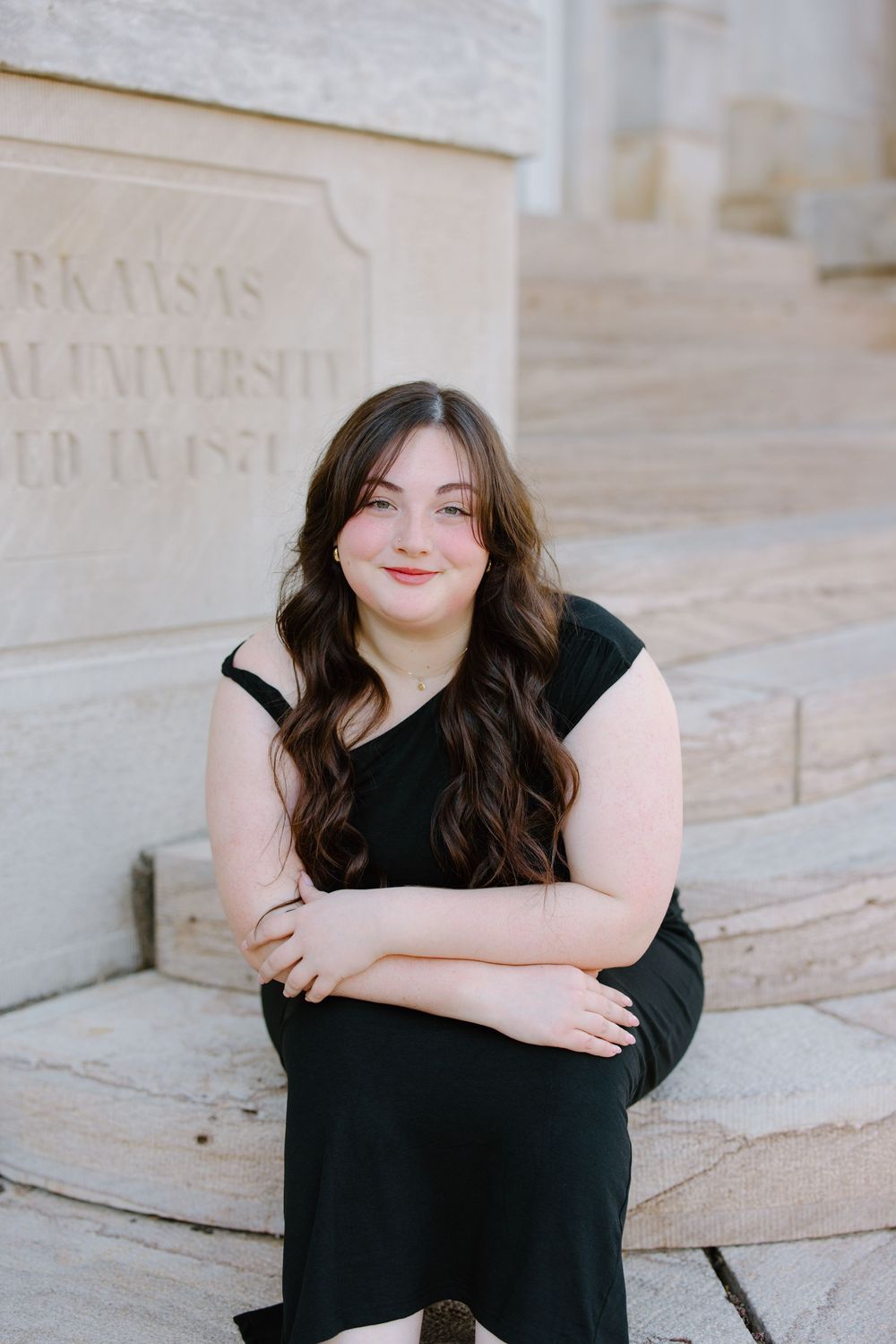 Young woman in black dress sitting on stone steps at Arkansas A&M University, smiling with long wavy brown hair.