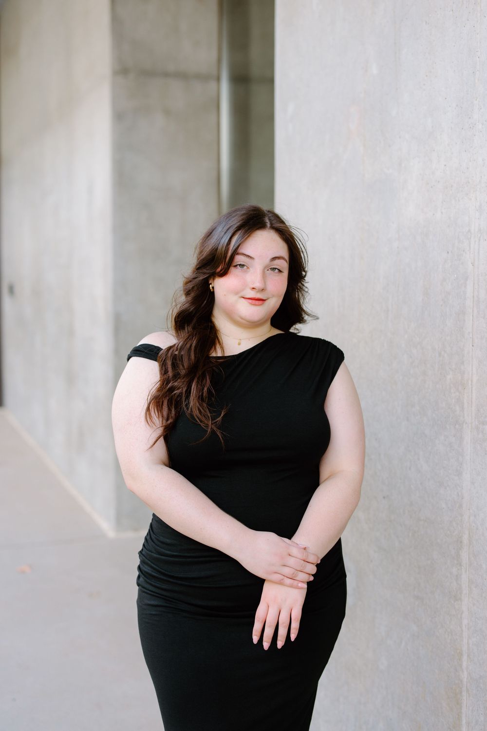 Young woman in black fitted dress posing confidently outdoors near stone columns, with long wavy brown hair.