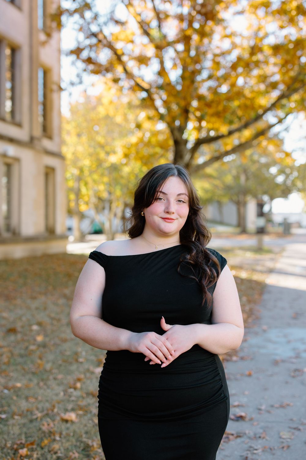 Young woman in black off-shoulder dress posing outdoors near autumn trees and a brick building.