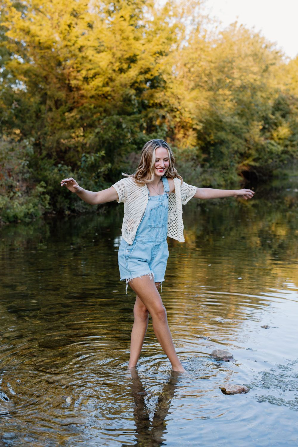 Person in overalls plays in creek water with autumn trees in background.