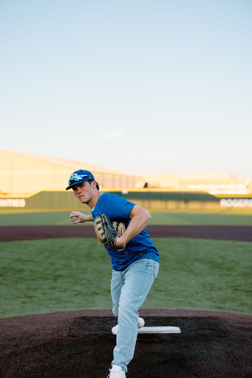 Baseball player in blue jersey and jeans practices pitching form on dirt mound at sunset on baseball field.