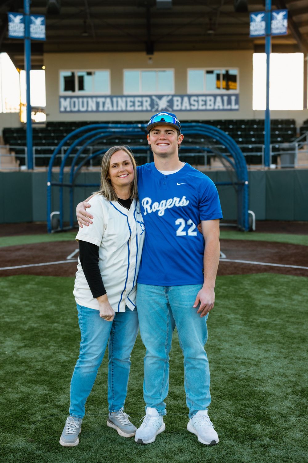 Baseball player wearing number 22 Rogers jersey poses on field at Mountaineer Baseball stadium with supporter.
