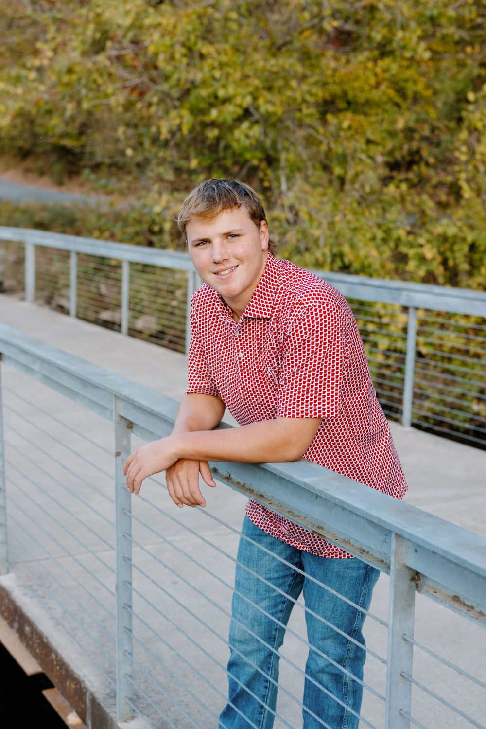 A person in a red and white checkered shirt leans against a railing on a wooden walkway surrounded by foliage.