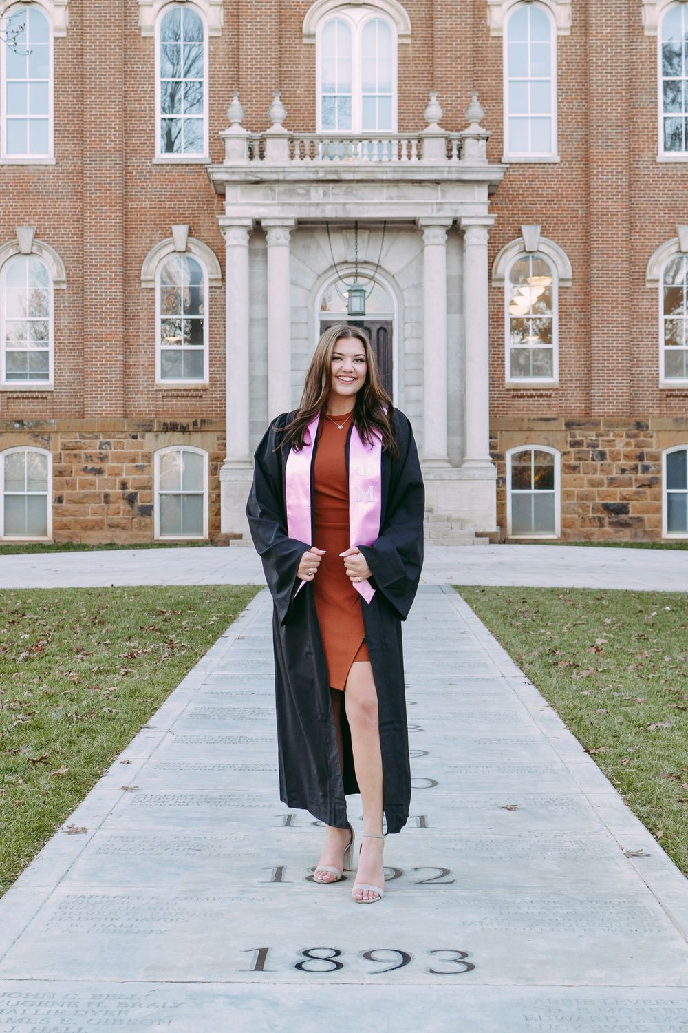 Graduate in rust dress and black graduation gown poses in front of historic brick building.