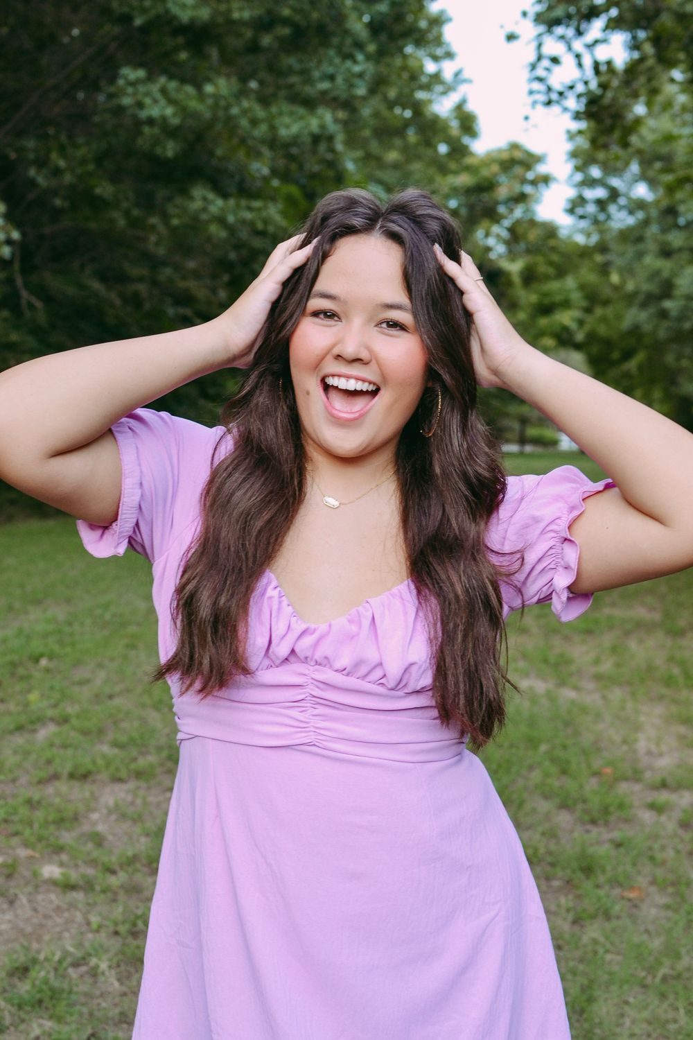 Person in pink dress laughing with hands in hair outdoors on sunny day.