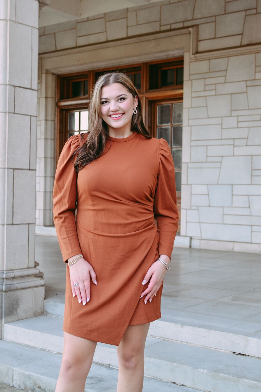 Young woman in a stylish rust-colored dress with puff sleeves poses on stone steps.
