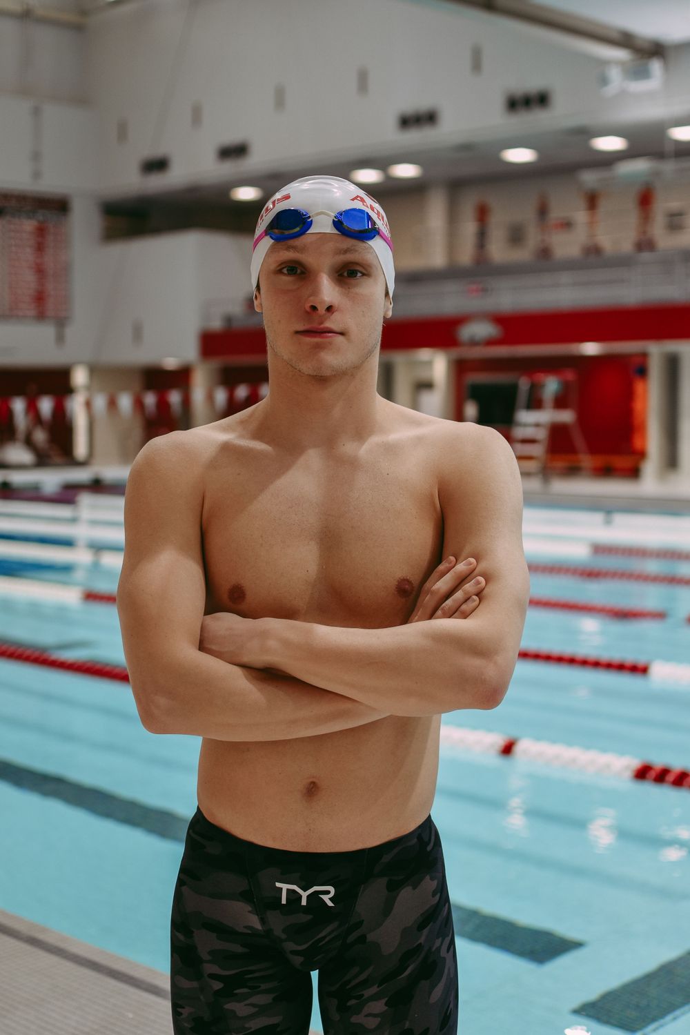 Competitive swimmer stands by pool wearing swim cap and goggles with arms crossed.