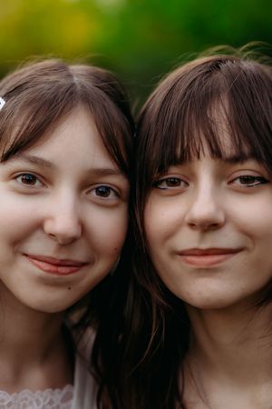 Two young women with brown hair and bangs smile closely together outdoors with green bokeh background.