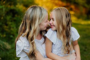 Two blonde girls in white dresses share a joyful moment, smiling and whispering in a sunlit outdoor setting.