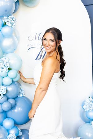 Smiling bride in white dress posing beside blue balloon arch with hydrangeas at bridal shower.
