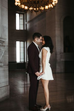 Couple sharing a romantic moment in an elegant hall, man in burgundy suit and woman in white mini dress under a chandelier.