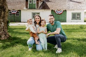 Happy family of four posing outdoors in front of a green barn decorated with patriotic bunting on a sunny day.