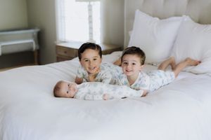 Two older siblings smile with newborn baby lying on white bed in bright bedroom.