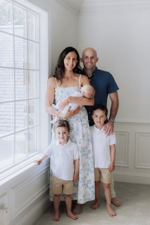 Family of five posing indoors, parents with three children including a newborn, dressed in coordinating blue and white outfits.