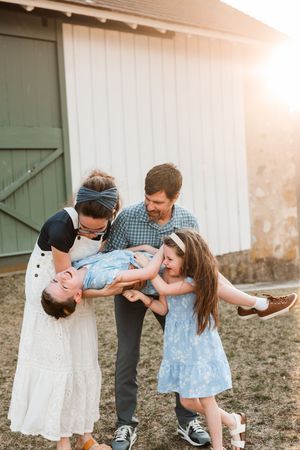 A joyful family of five plays together outdoors at golden hour near a rustic barn, with parents lifting a laughing child.