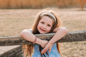 A smiling young girl with long hair and a headband leans on a wooden fence in a sunny outdoor setting.