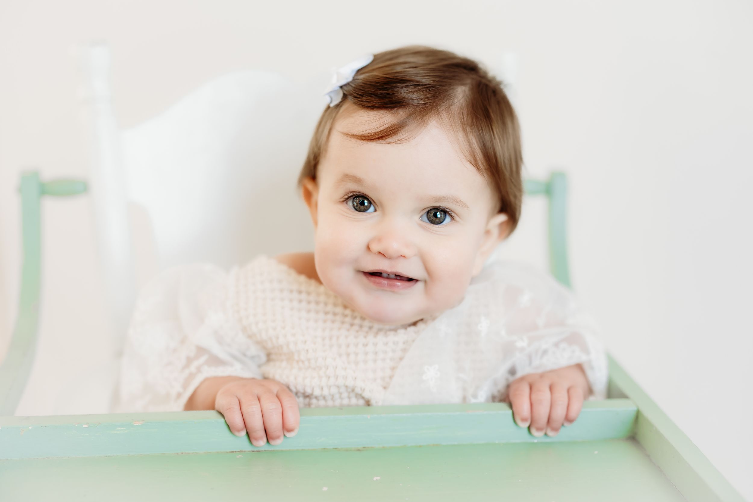 Smiling baby with brown hair sitting in a mint green high chair, wearing a white lace outfit against a light background.