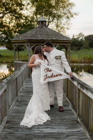 Newlyweds kiss on a wooden dock holding a pennant flag reading The Jacksons, with a gazebo and pond backdrop.