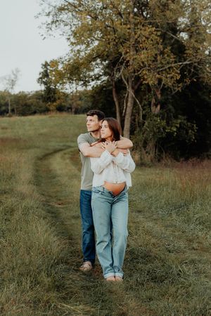 A couple embraces in a green field, man hugging woman from behind, both in jeans, with trees in background.