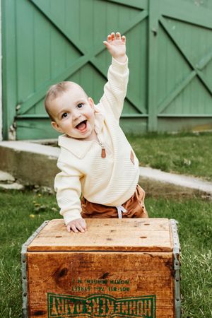Happy baby in cream sweater raising hand while leaning on vintage wooden crate outdoors near green barn door.