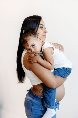 Pregnant woman in white crop top and jeans holds young child on her hip against a light background.