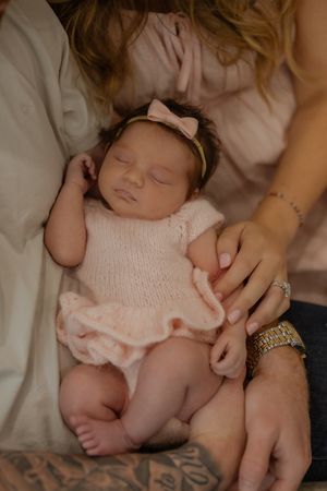 Sleeping newborn baby girl in pink knit dress and bow headband cradled in parents arms.