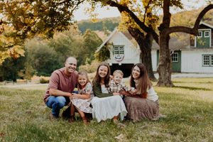 Family of five smiling outdoors on grass near a white barn and large tree during golden autumn afternoon.
