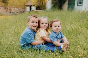 Three young siblings sitting together in a green meadow with wildflowers, smiling outdoors near a historic building.